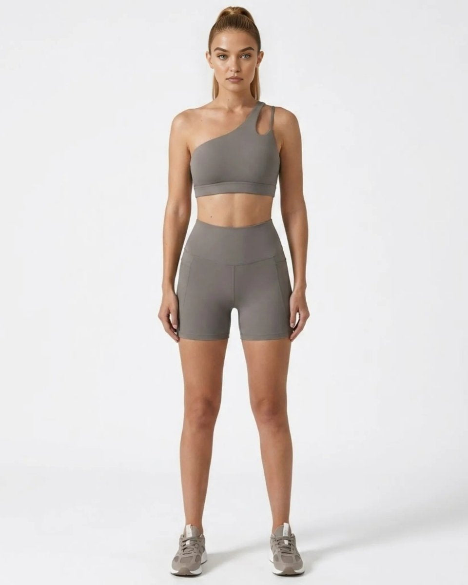 Front view of woman wearing grey asymmetrical one shoulder sports bra and high waisted biker shorts activewear set.