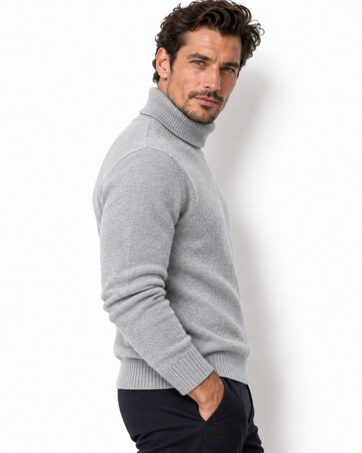 A man wearing a light gray ribbed turtleneck sweater and dark trousers, posing in profile against a clean white background.