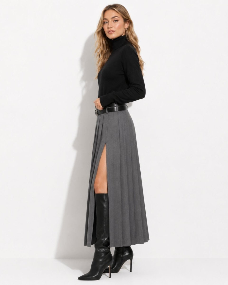 A woman models a long gray pleated midi skirt with a high side slit, styled with a black belt, a black turtleneck top, and black knee-high boots against a plain white studio background.