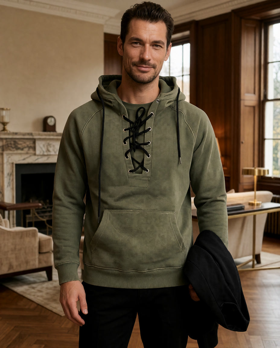 Ein Model trägt den olivgrünen Cannon Kapuzenpullover in einem Raum mit Kamin und hält eine schwarze Jacke.