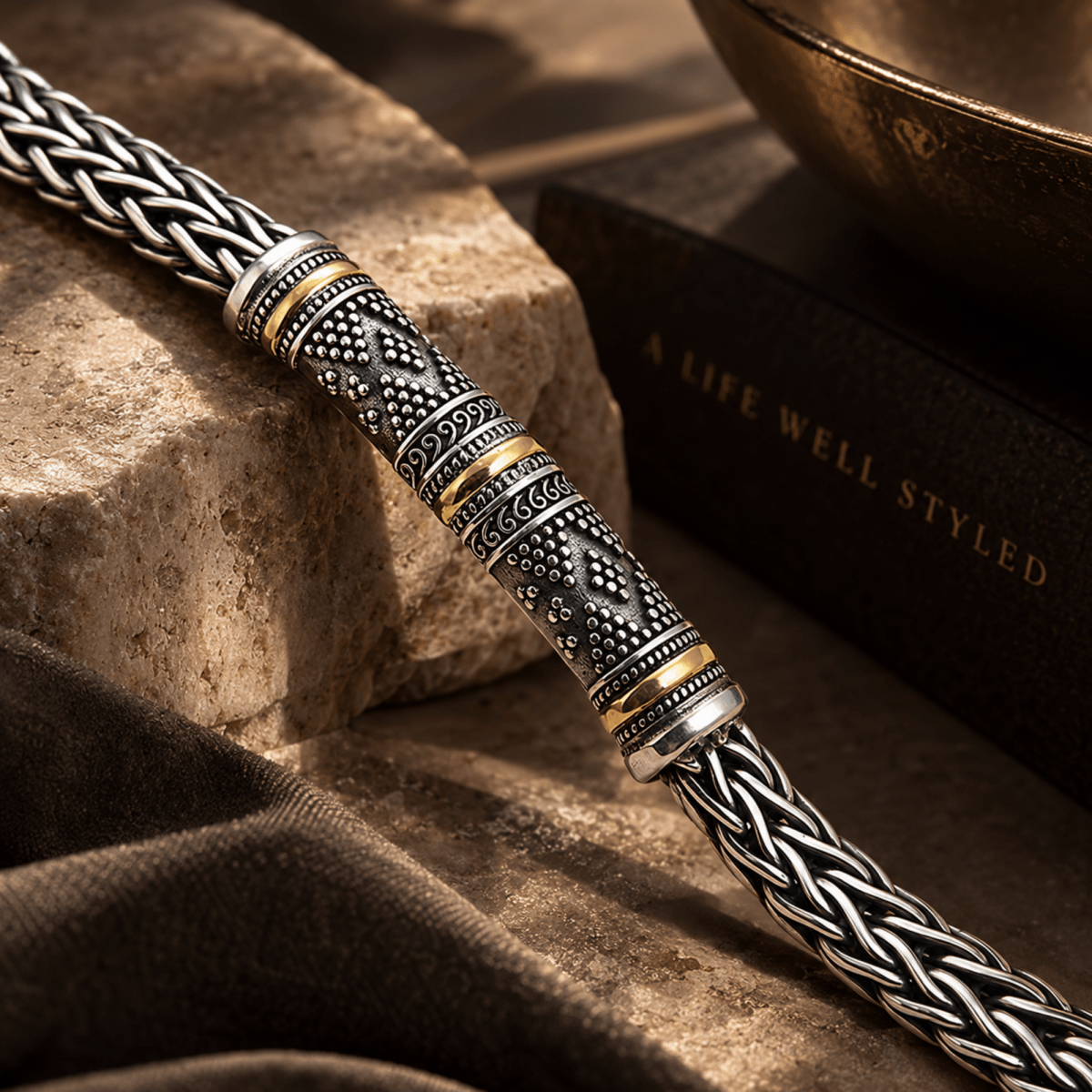 A close-up of a braided metal bracelet with an ornate cylindrical connector featuring engraved geometric patterns and gold-tone accents, styled on draped fabric and stone with a dark book in the background.