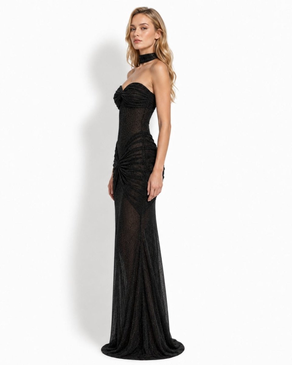 A woman modeling a strapless black evening gown with a high neck, ruched bodice, sheer mesh panels, and a floor-length silhouette against a plain light background.