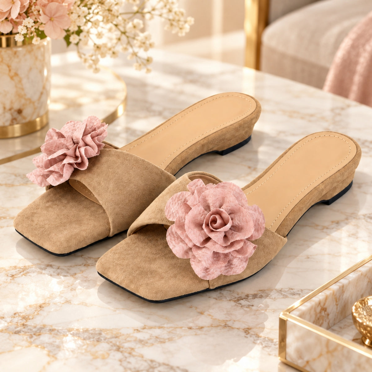 A pair of khaki suede mule sandals with large pink fabric flowers on the toe, displayed on a marble table in a styled interior setting.