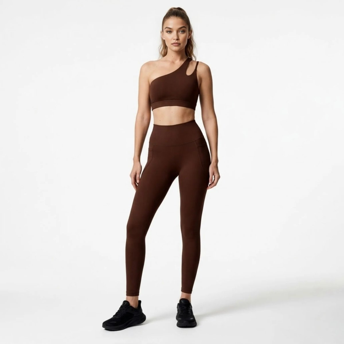 Front view of female model wearing Kouvr dark brown one shoulder activewear set featuring an asymmetrical sports bra and high waisted leggings with pockets.