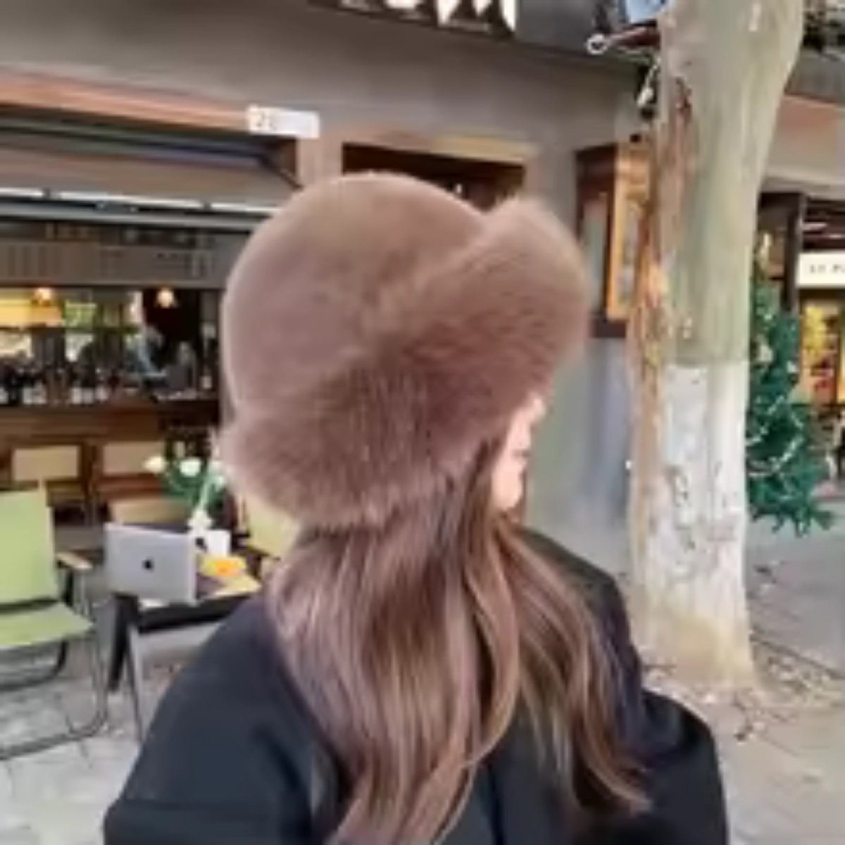 Person wearing a fluffy brown hat in an indoor setting with a Christmas tree in the background
