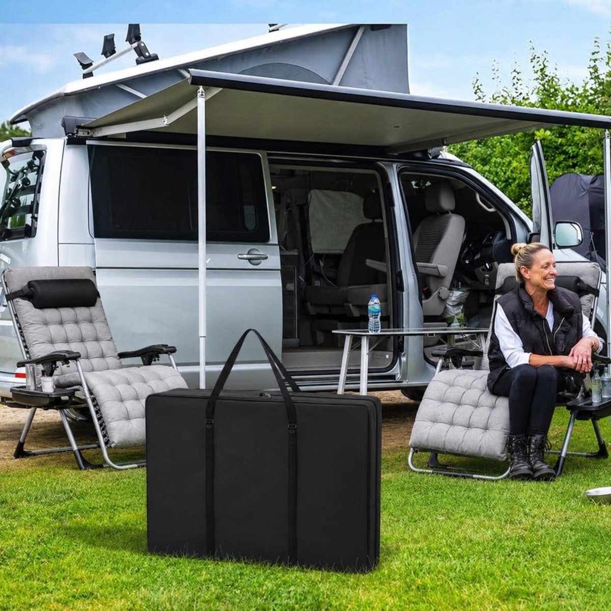 Black Kouvr heavy duty folding chair storage bag sitting on green grass next to a luxury travel RV and grey outdoor lounge chairs.