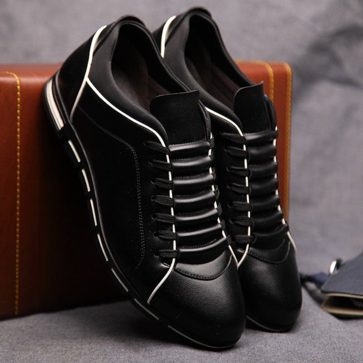 A pair of black leather Kouvr Jimmy men's sneakers with white trim piping and a ribbed lacing panel, propped against a brown suitcase.

