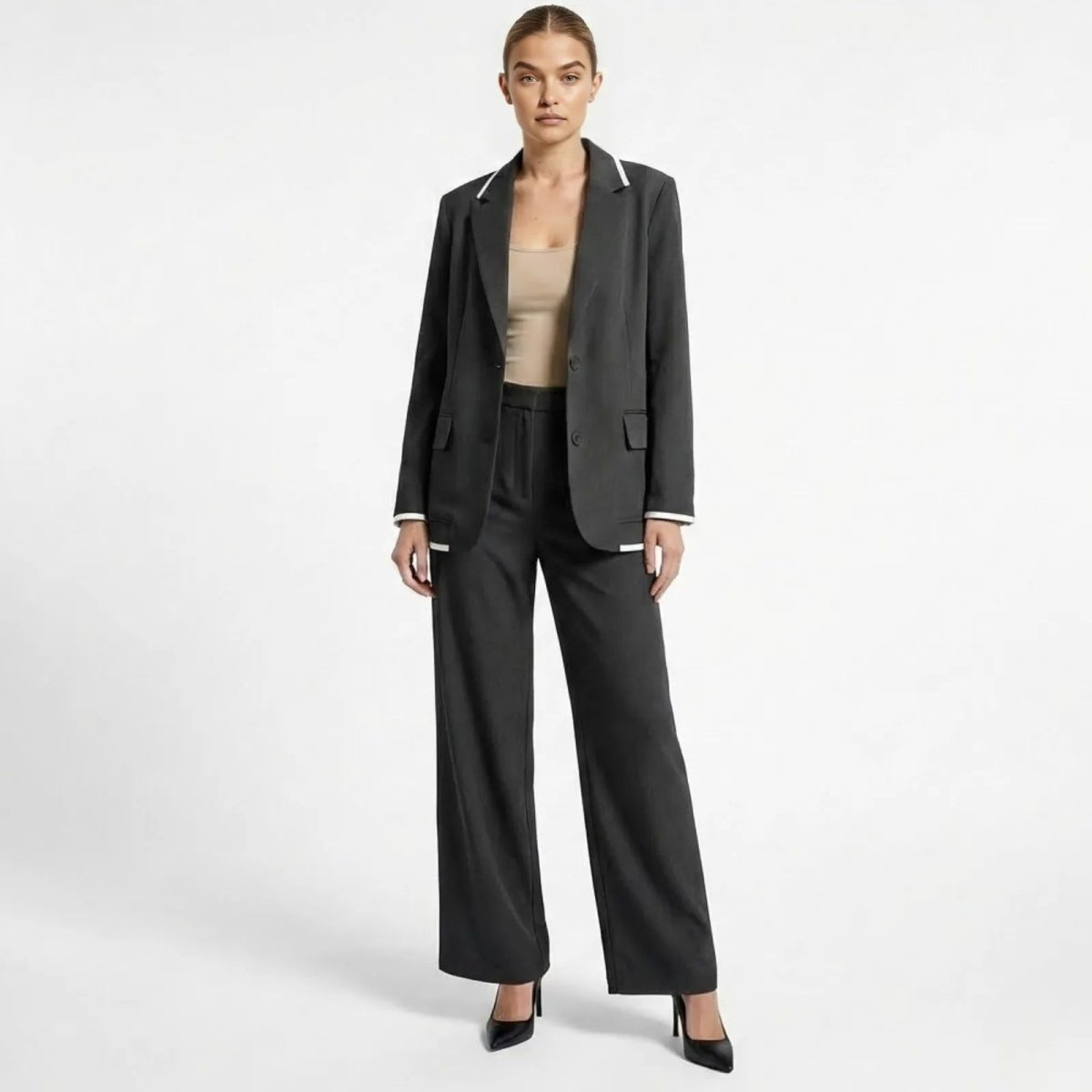 Model wearing the Kouvr Lucia Set two-piece pant suit in dark grey, featuring a single-breasted blazer with white trim on the lapels and matching wide-leg trousers, styled with a nude top and black heels. 