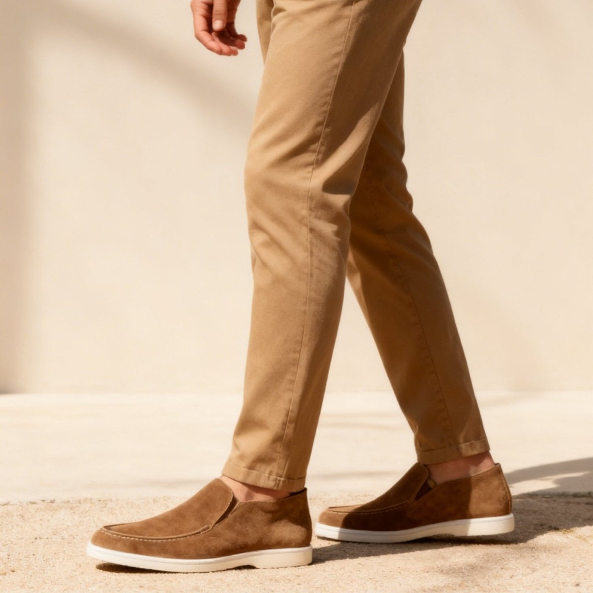 A man walking, showing the side profile of the brown suede loafer boot, paired with light brown trousers in natural sunlight.
 