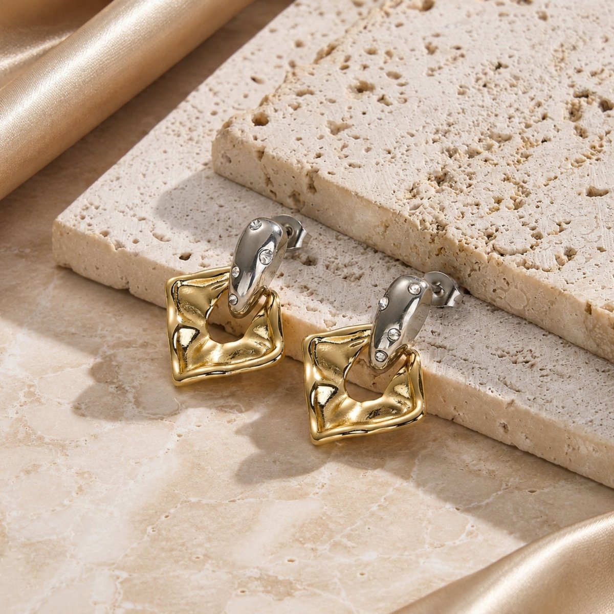 Kouvr abstract gold and silver dangle earrings displayed on textured beige travertine stone slabs bathed in warm sunlight.
