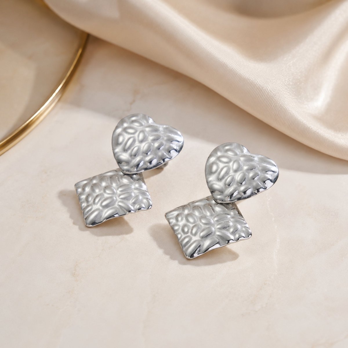 Close up detail of Kouvr silver hammered heart and geometric rhombus earrings showing premium metallic finish