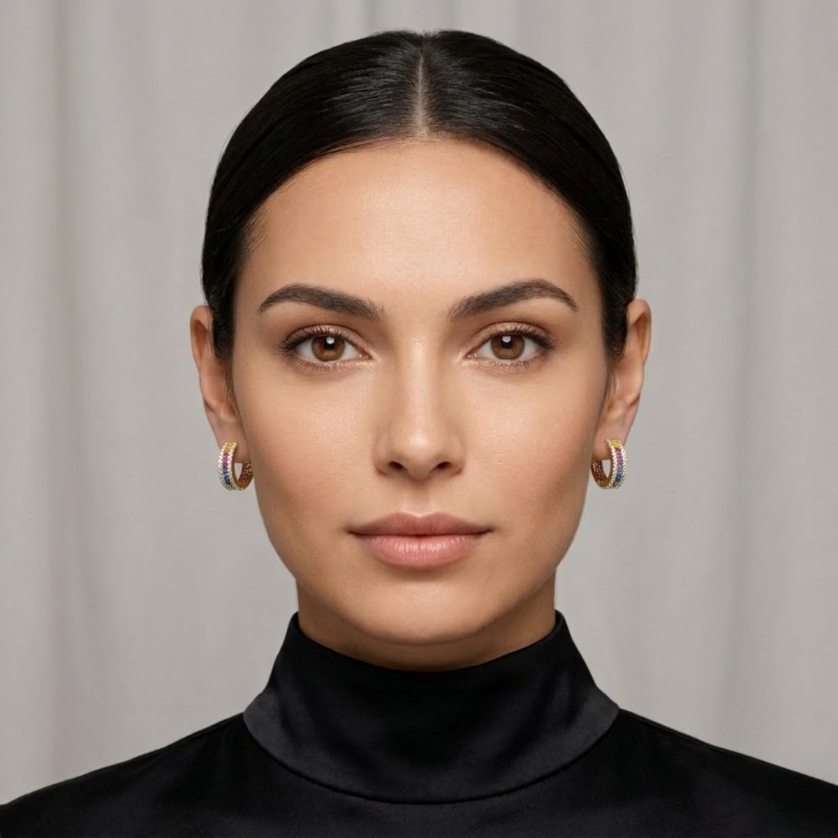 A close-up of a woman with dark hair pulled back, wearing the Kouvr Yasmina rainbow and diamond gold huggie hoop earrings, showcasing their size and scale.
