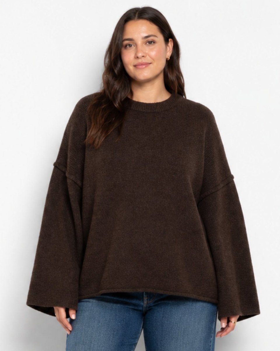 A woman wearing a dark brown oversized knit sweater with wide bell sleeves and blue jeans, standing against a plain light gray studio background.
