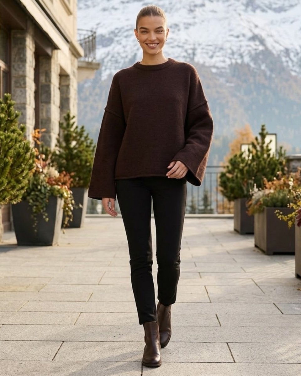 A woman walking outdoors on a stone walkway wearing a dark brown oversized knit sweater, black skinny pants, and brown ankle boots, with mountains and potted plants in the background.