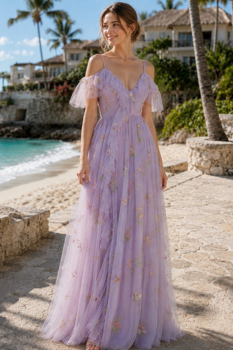 A woman wearing a lavender off-the-shoulder tulle maxi dress with gold floral embroidery, standing on a stone path near a tropical beach with palm trees and ocean in the background.