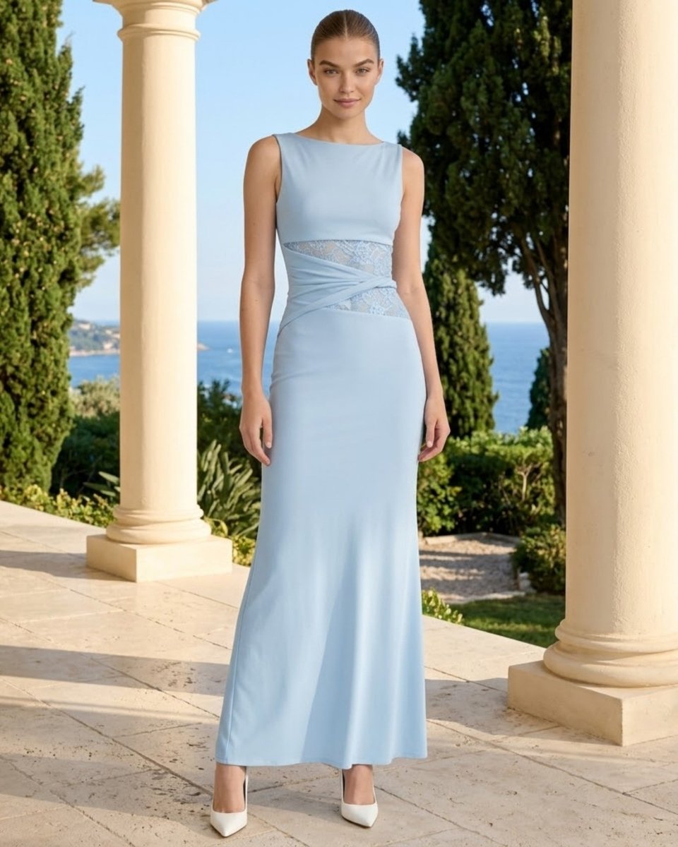 A woman stands outdoors between large cream columns, wearing a sleeveless light blue floor-length gown with a sheer lace waist detail and white pointed shoes. Trees and blue water are visible in the background.