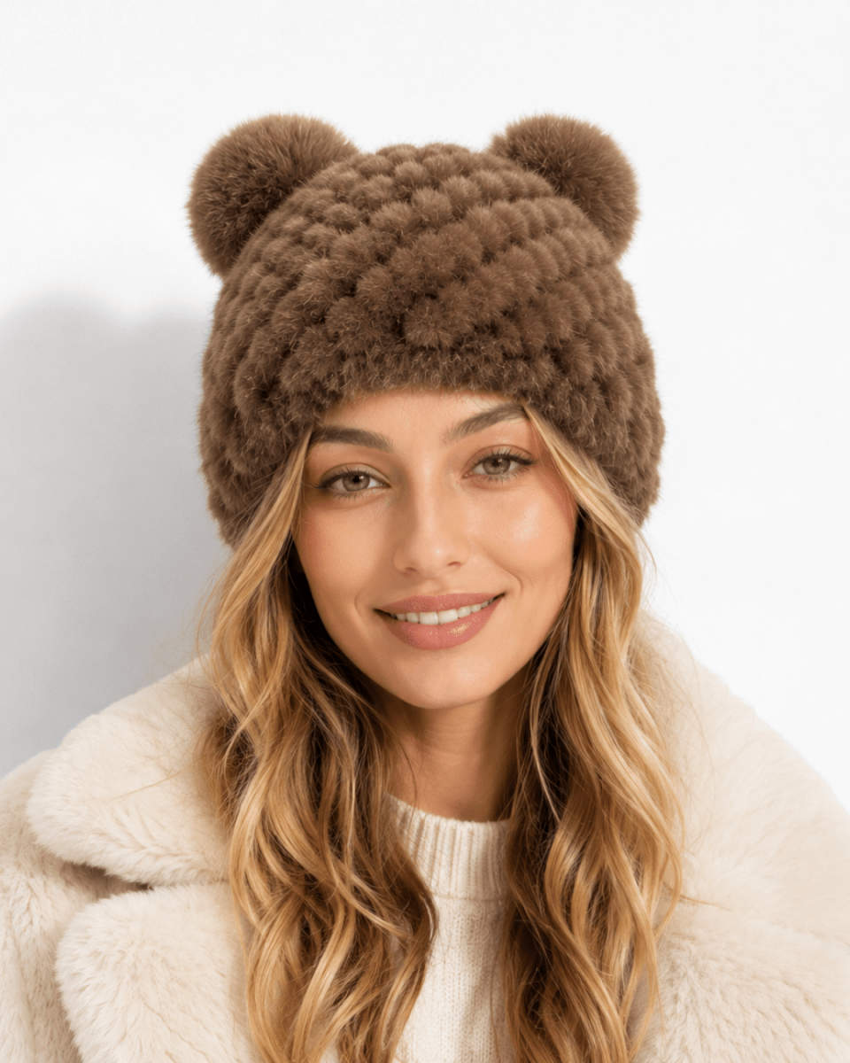 A woman wearing a brown chunky knit beanie with two pom-poms on top, paired with a cream faux-fur collar, against a clean white background.