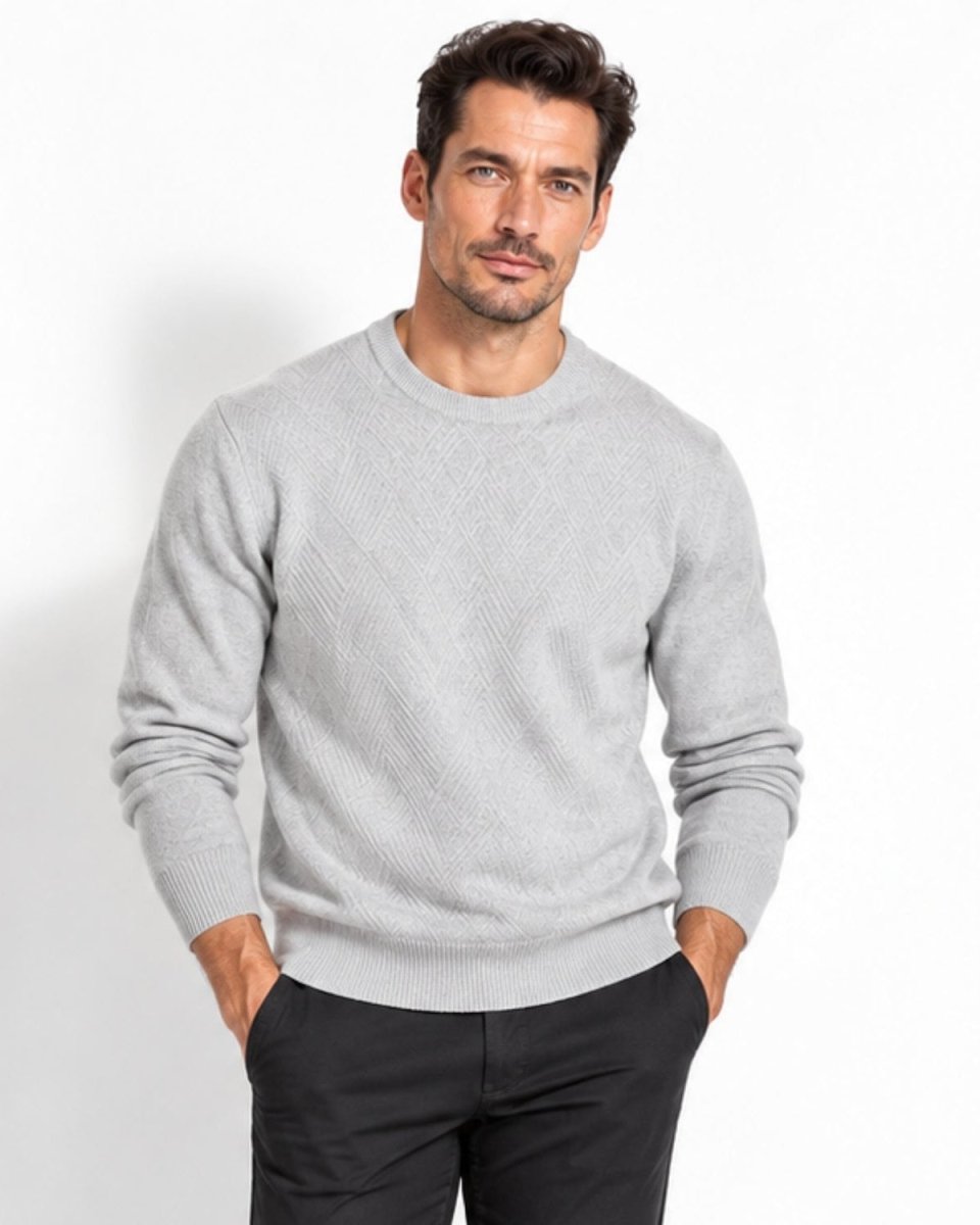 A man wearing a light gray textured knit crewneck sweater and dark trousers, standing against a plain white studio background.