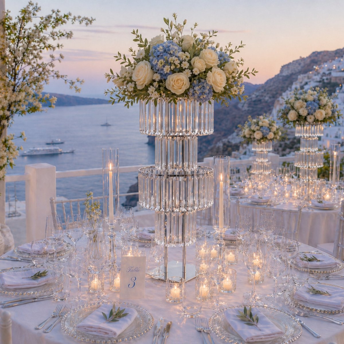 Tall tiered crystal wedding centerpiece stand decorated with white and blue flowers on a banquet table overlooking an ocean sunset.
