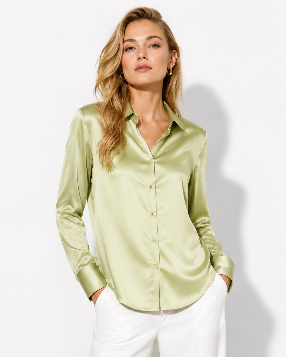 A woman modeling a light olive green satin button-down shirt with white pants, standing against a plain white studio background.
