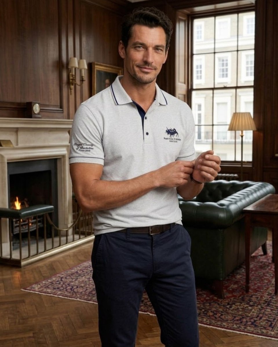 A man wearing a light gray polo shirt with a navy blue collar stripe and embroidered logo, paired with navy pants, standing in a classic room with wood paneling, a fireplace, and a green leather sofa.