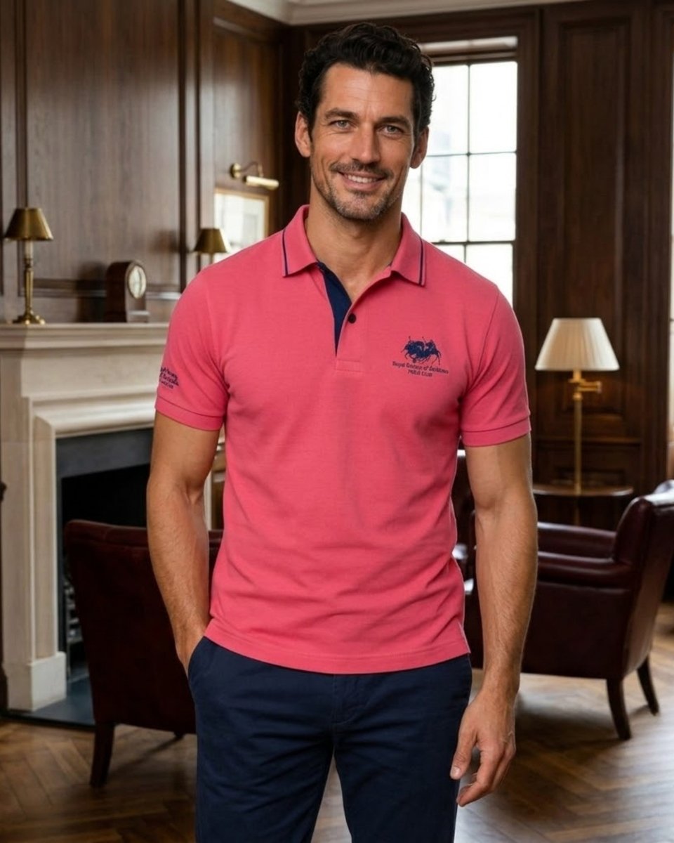 A man wearing a coral pink polo shirt with a navy blue collar and chest logo, paired with navy pants, standing in a classic, elegant room with wood paneling and a fireplace.