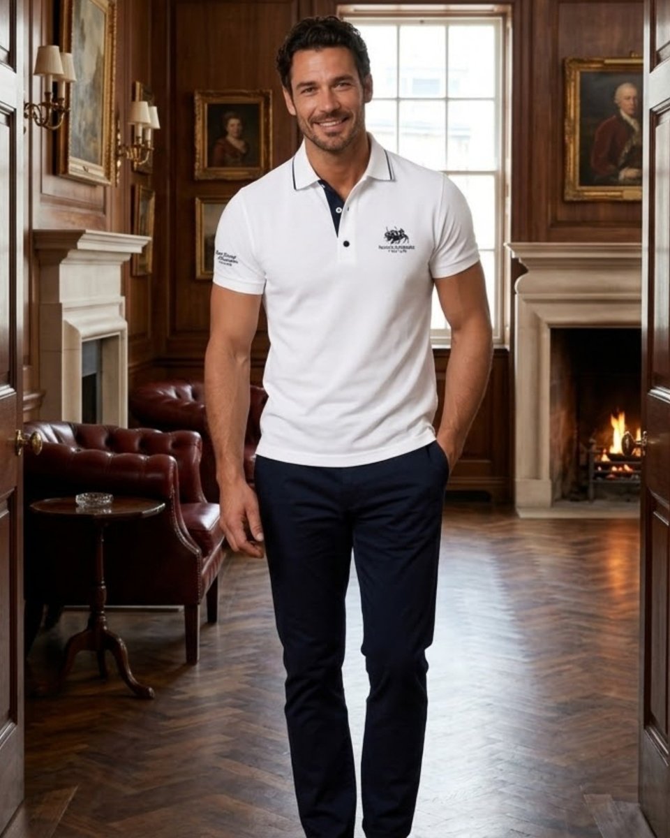 A man wearing a white short-sleeve polo shirt with a navy collar and a small embroidered logo on the left chest, paired with dark navy trousers, standing in a classic, wood-paneled room with fireplaces.