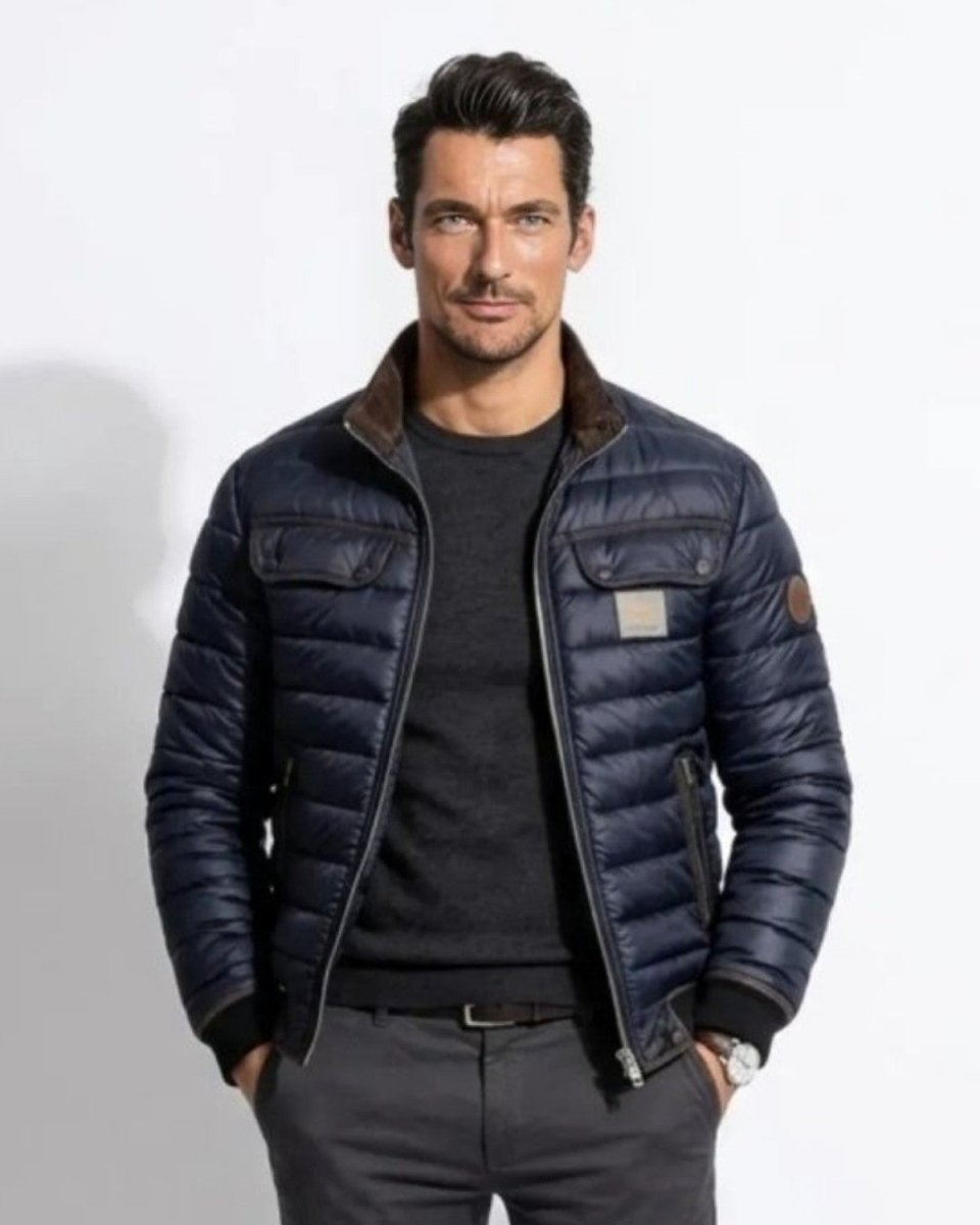 A man wearing a navy blue puffer jacket with brown collar lining, a dark gray crewneck sweater, and dark gray trousers, standing against a white studio background.