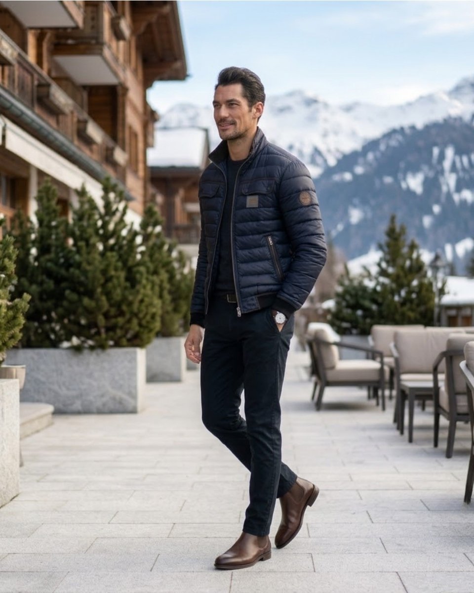 A man wearing a dark navy quilted puffer jacket, dark trousers, and brown leather boots walks along a paved outdoor path in a mountain resort setting with wooden chalets and snowy peaks in the background.