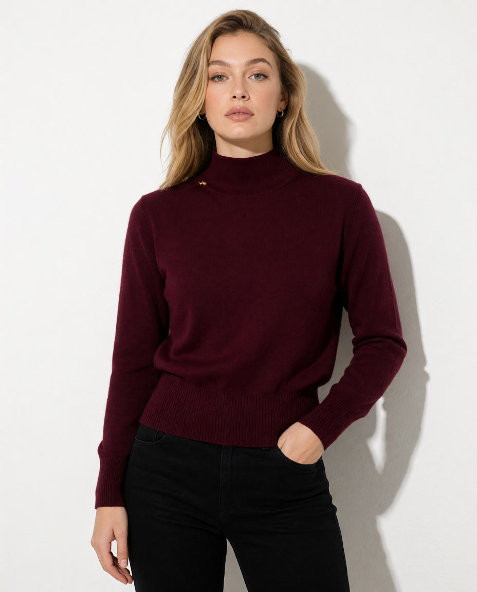 A woman modeling a long-sleeve ribbed mock neck sweater in deep burgundy, paired with black pants, against a plain white wall.