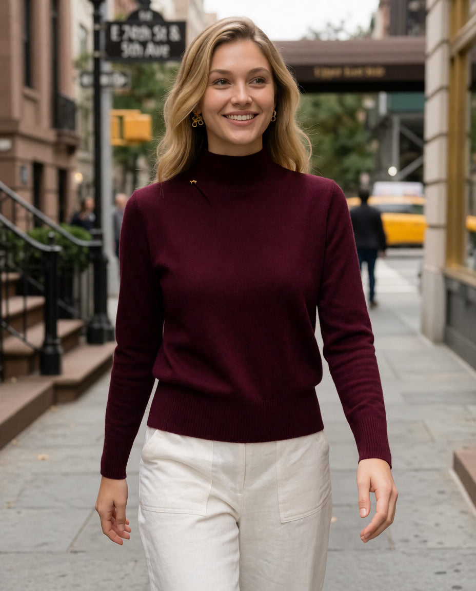 A woman walking on a city street wearing a fitted burgundy mock-neck knit sweater and white pants.