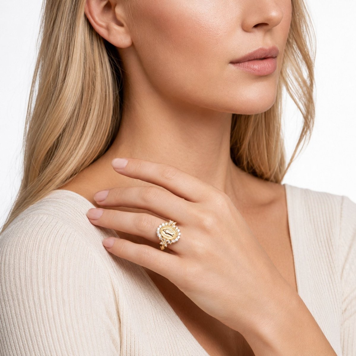 A close-up lifestyle shot of a woman wearing a gold ring with a floral or sunburst design set with small clear stones on her ring finger. She is wearing a cream-colored ribbed top against a clean white background.