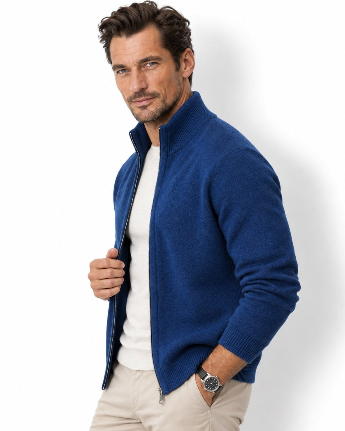 A man wearing a navy blue zip-up knit sweater over a white t-shirt and beige trousers, standing against a white studio background.
