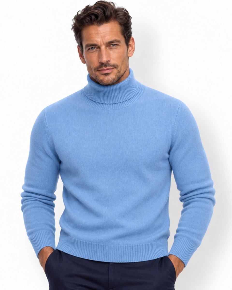 A man wearing a light navy knit turtleneck sweater and dark navy trousers, standing against a plain white studio background.