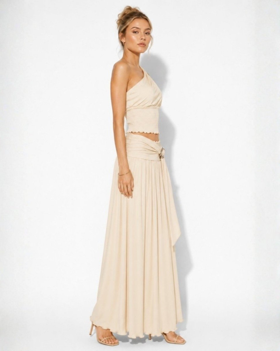 A woman modeling a two-piece beige dress set consisting of a halter crop top and a long pleated skirt, photographed against a plain white studio background.