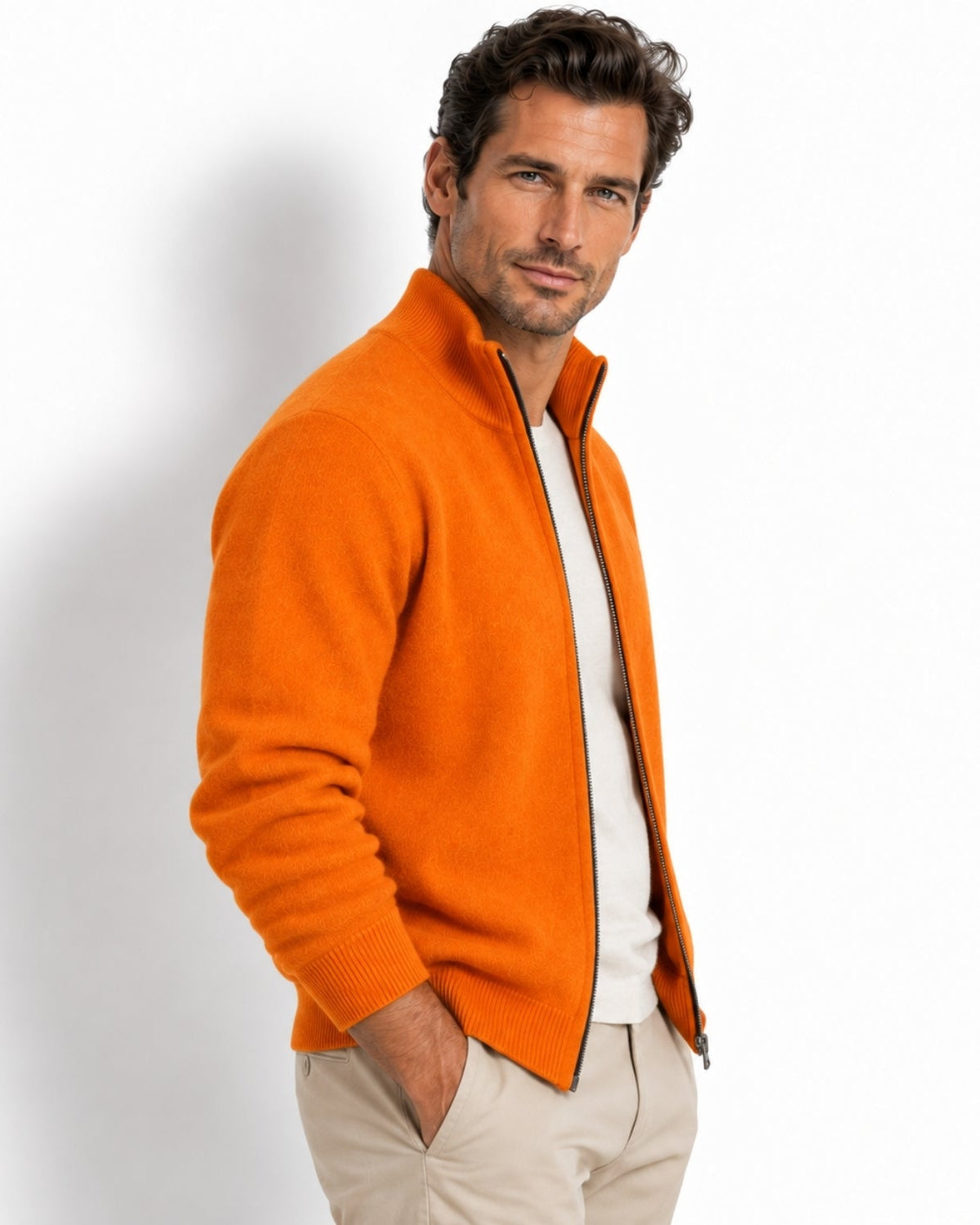 A man wearing a bright orange knit zip-up sweater over a white shirt and beige trousers, standing against a plain white studio background.