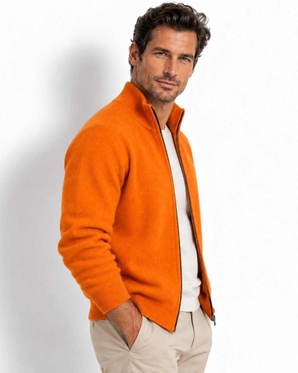 A man wearing a bright orange knit zip-up sweater over a white shirt and beige trousers, standing against a plain white studio background.