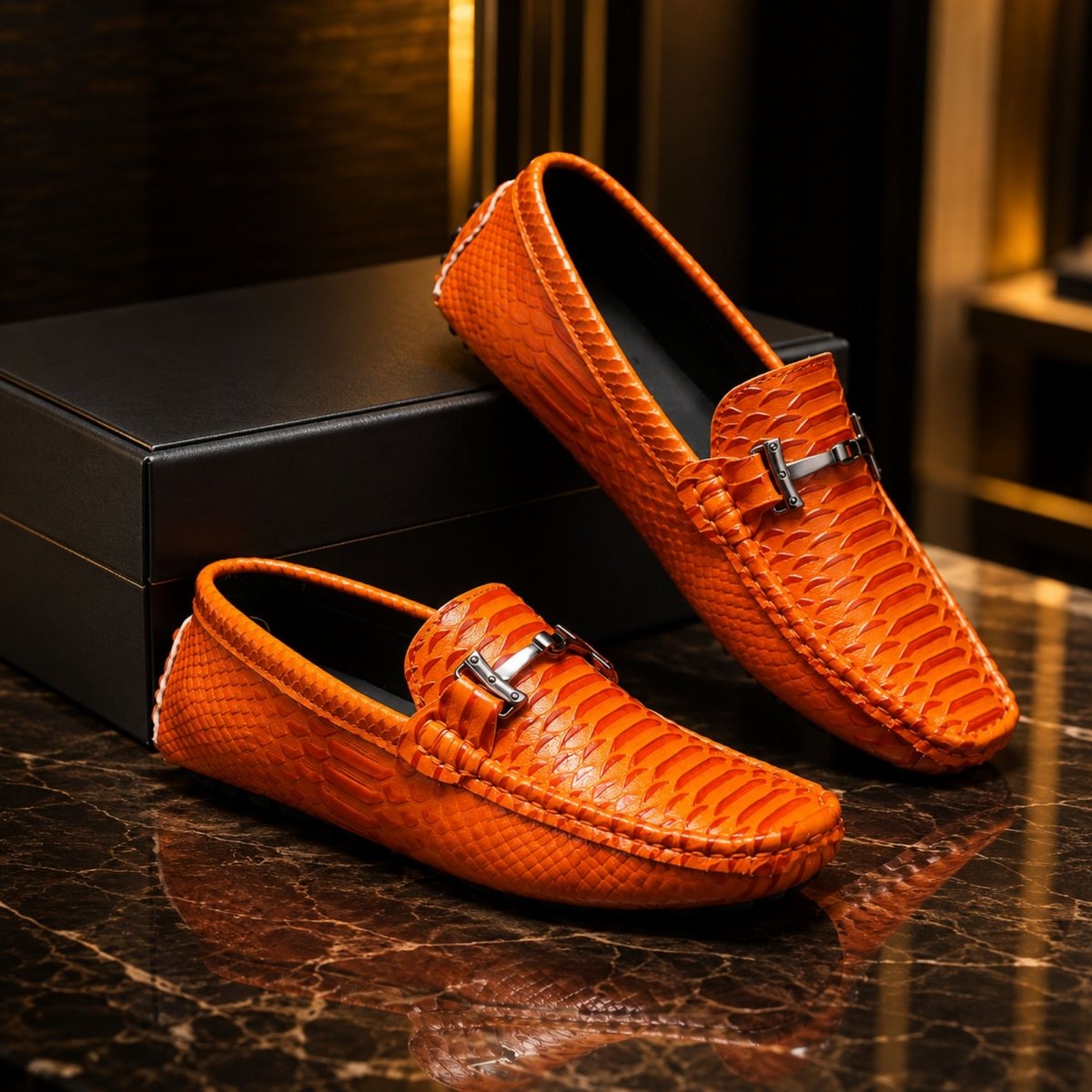A pair of orange textured snakeskin loafers with silver horsebit-style buckles, displayed on a dark marble surface next to a black shoebox.