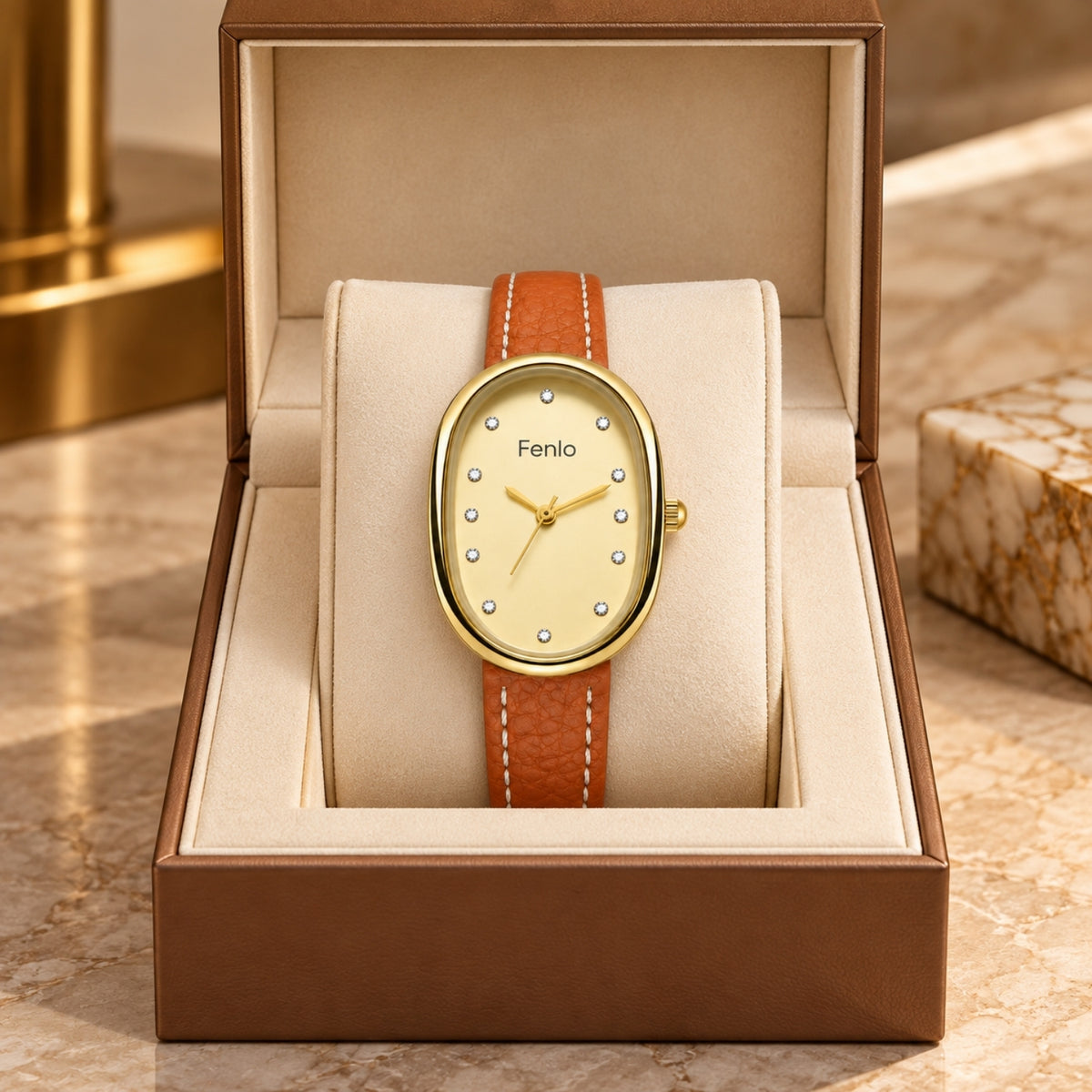 An oval wristwatch with a gold-tone case, cream dial, and orange-brown leather strap, displayed inside an open brown presentation box on a beige cushion. The dial shows the brand name 'Fenlo'.