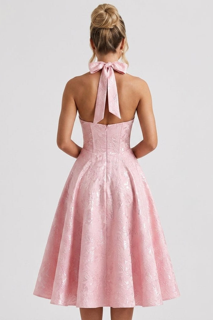 Back view of pink open back formal event dress highlighting large tied halter neck bow and pleated tea-length skirt.