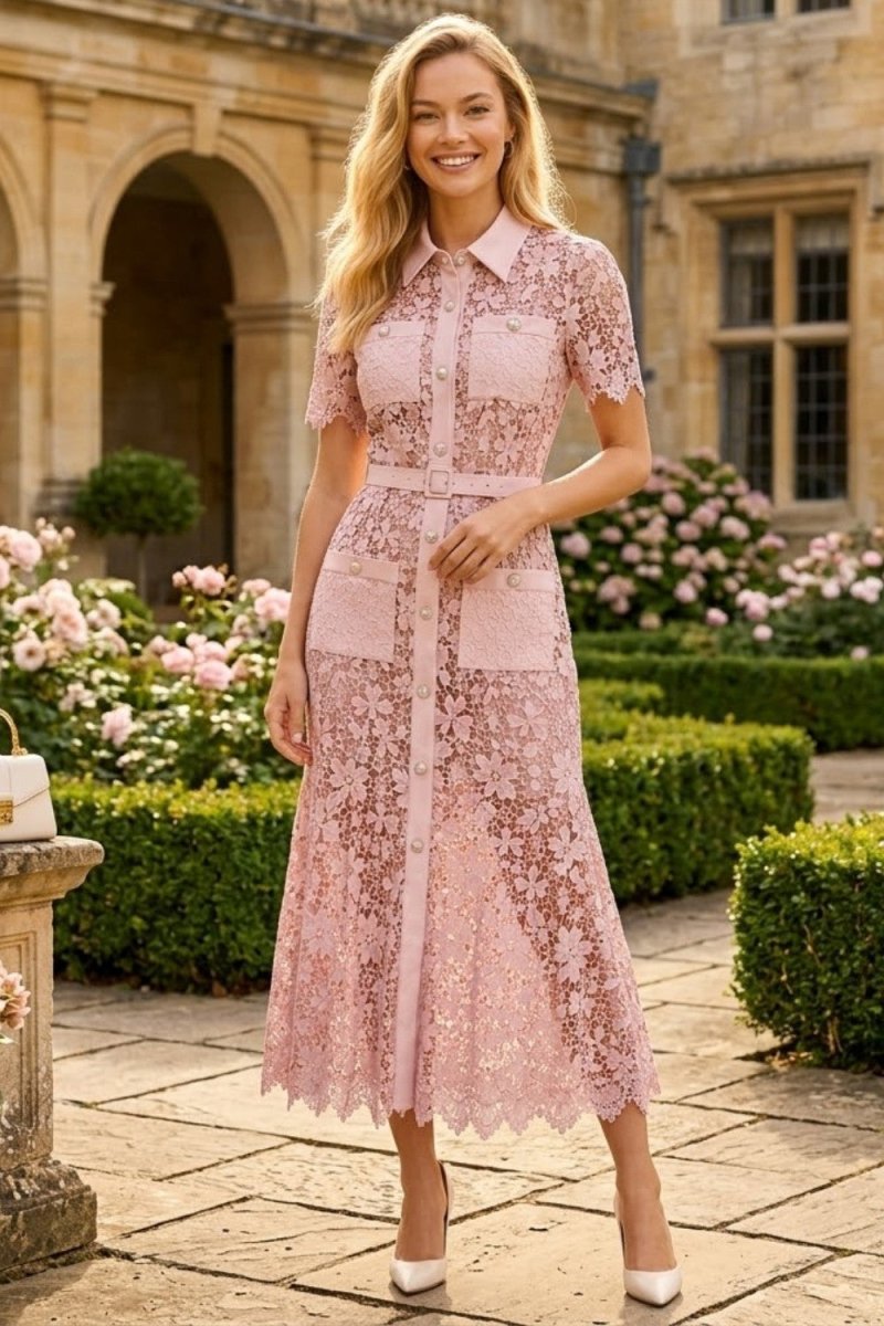 A woman stands in a garden courtyard wearing a light pink lace midi dress with short sleeves, a collared neckline, a self-tie waist belt, and a front slit. The background shows stone architecture, trimmed hedges, and pink flowers.