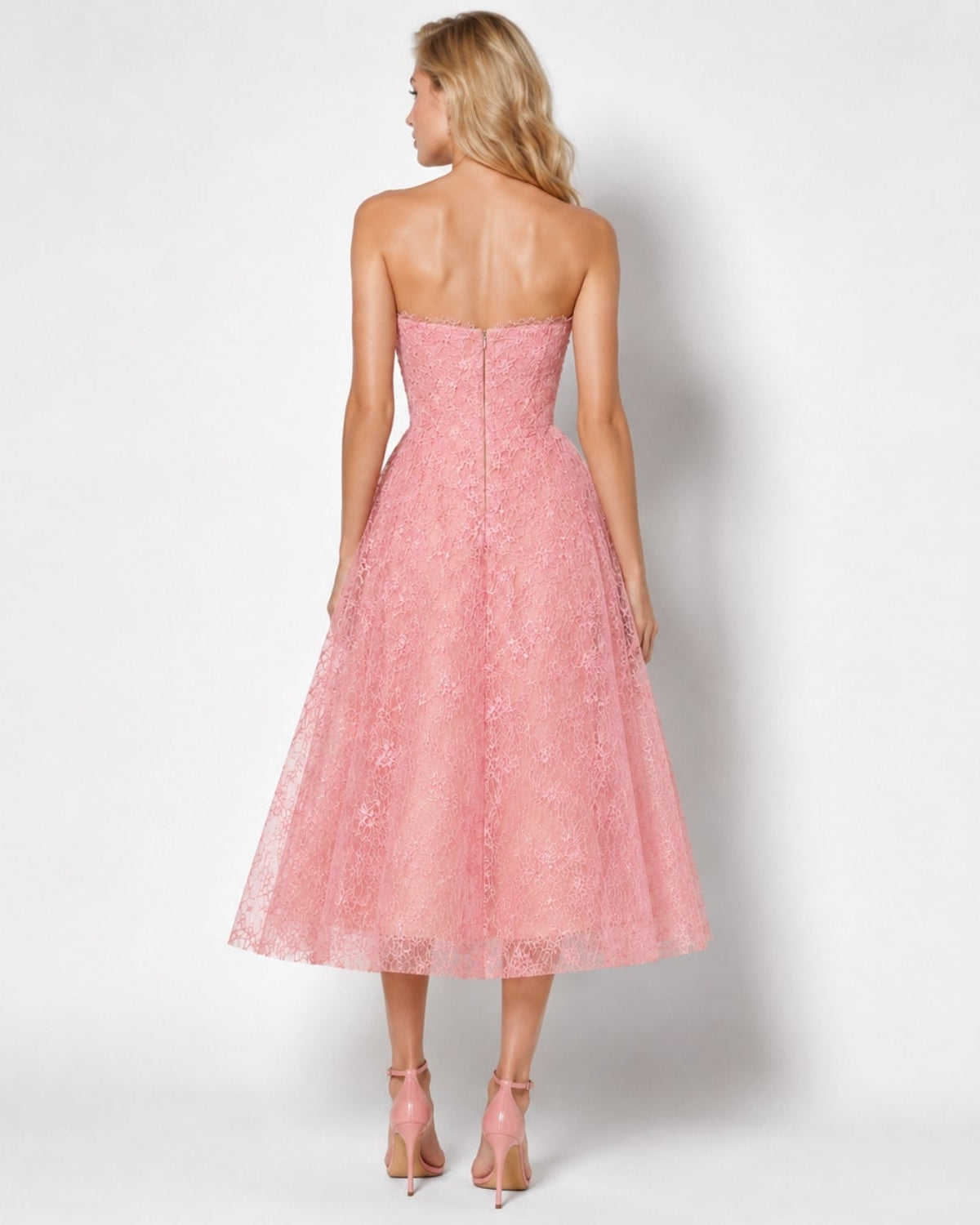A woman shown from the back wearing a strapless pink lace midi dress with a fitted bodice and flared skirt, paired with nude high heels, against a light gray studio background.
