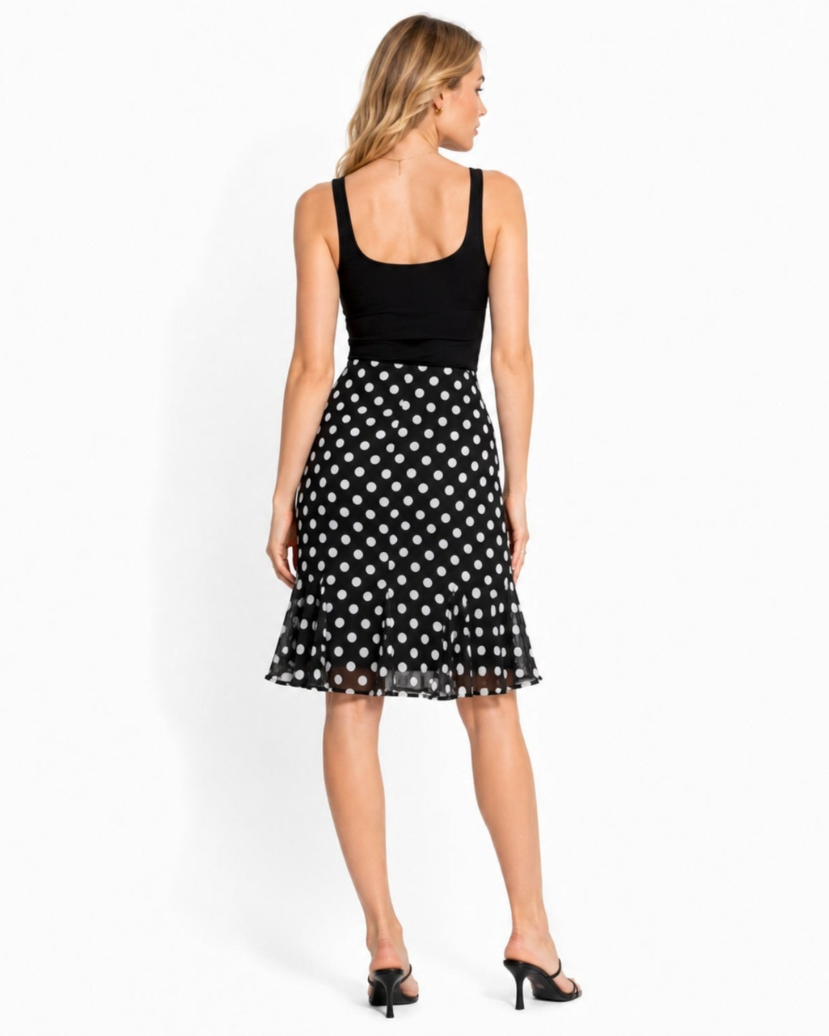 A woman shown from the back wearing a sleeveless black top and a black midi skirt with a white polka dot pattern, paired with black mule heels, against a plain white studio background.