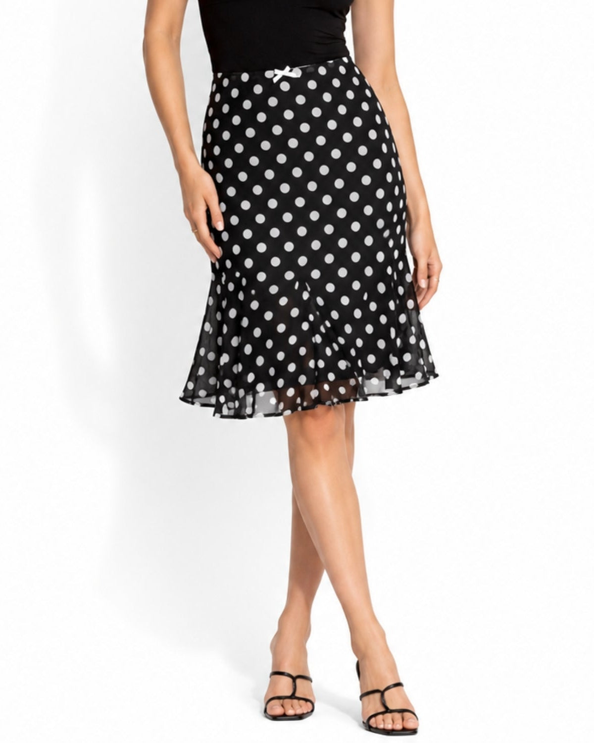 A woman models a black midi skirt with a white polka dot pattern, paired with a black top and black strappy sandals, against a plain white studio background.