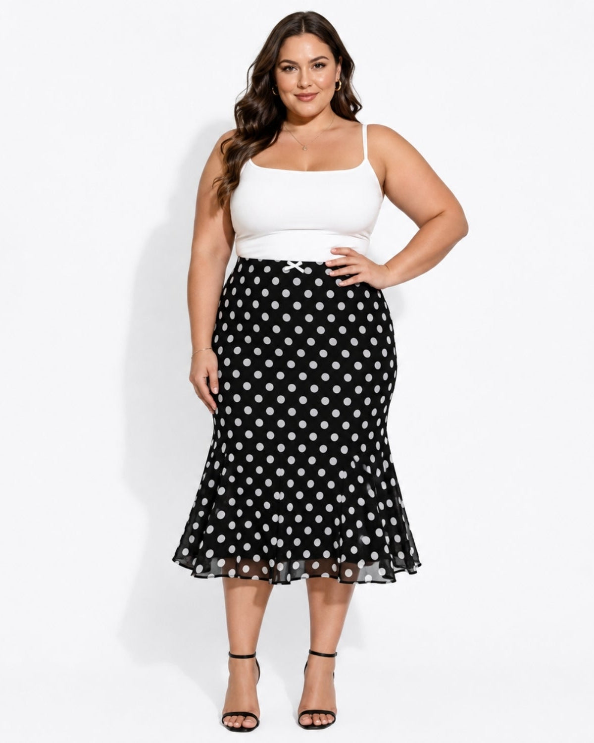 A woman modeling a black midi skirt with a white polka dot pattern, paired with a white spaghetti-strap top and black high-heeled sandals, against a plain white studio background.