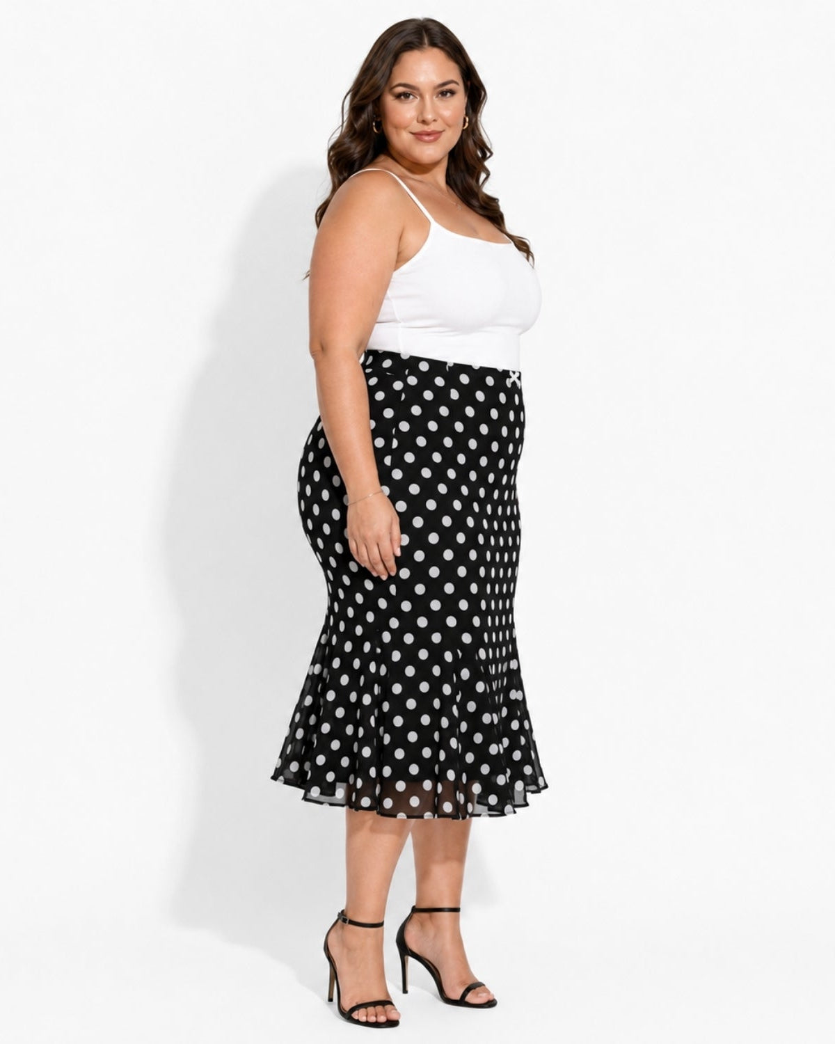 A woman modeling a black midi skirt with a white polka dot pattern, styled with a white camisole top and black strappy heels against a plain white studio background.