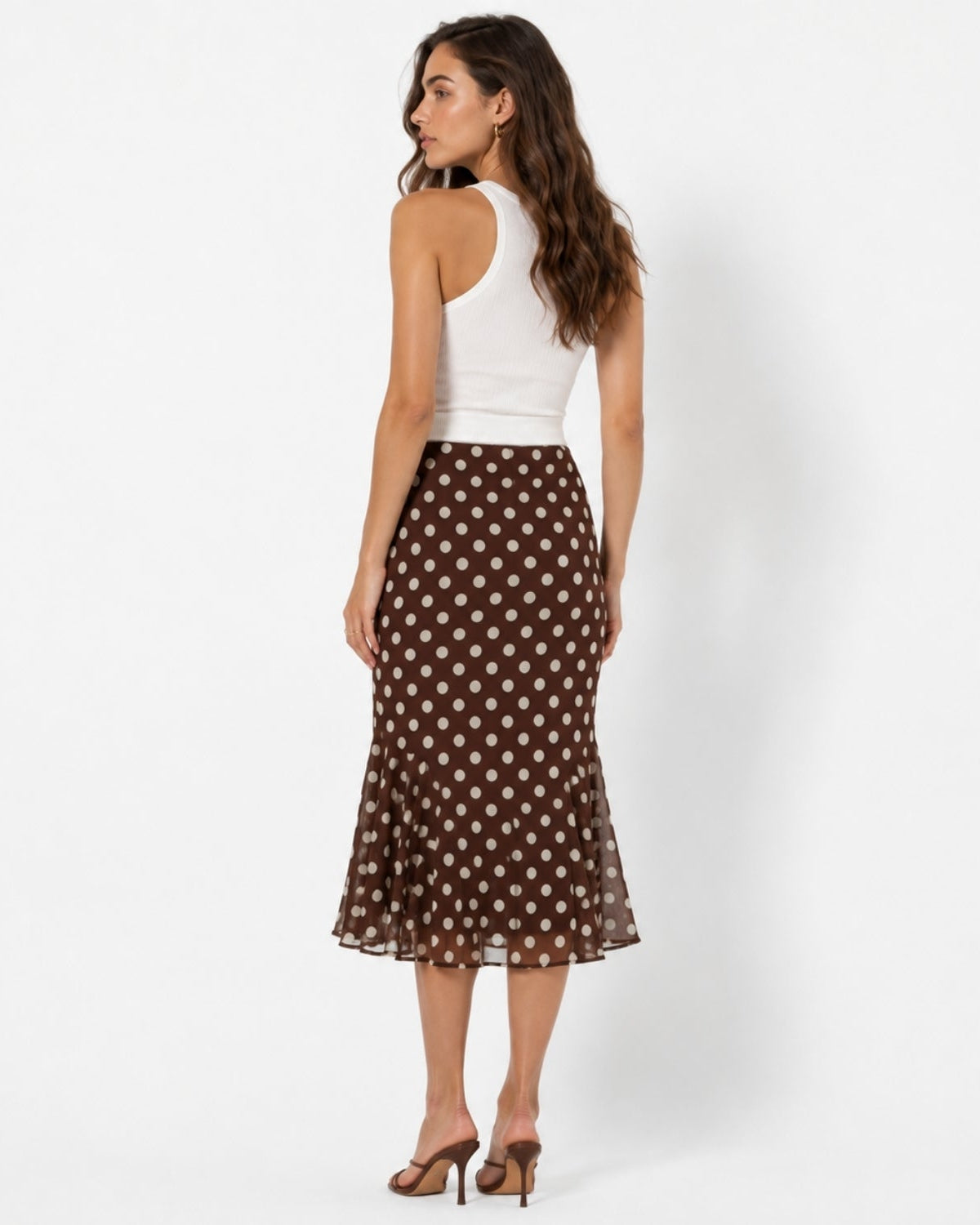 A woman stands in profile wearing a white ribbed tank top and a brown midi skirt with a beige polka dot pattern, paired with brown heeled sandals, against a plain white studio background.