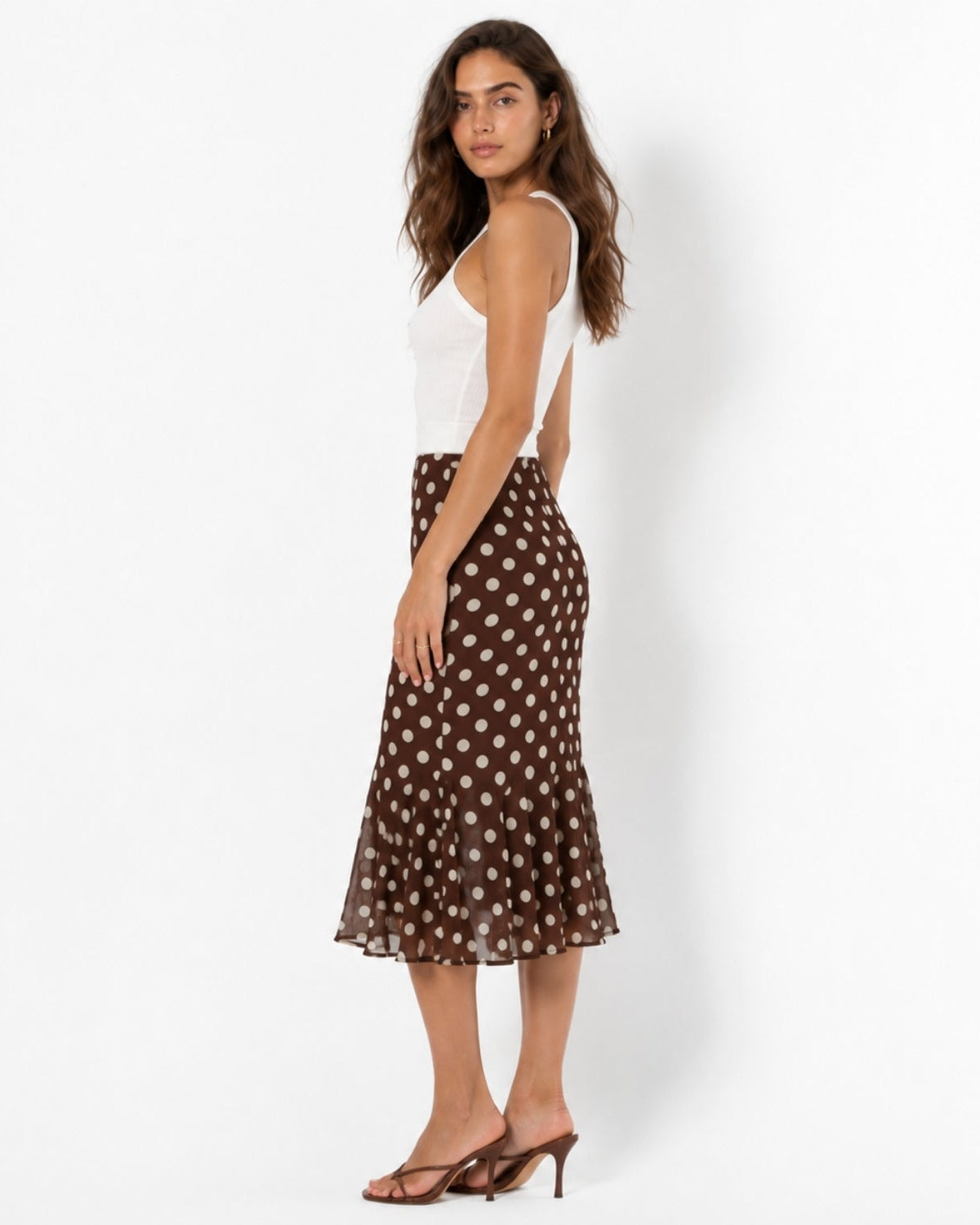 A woman modeling a brown midi skirt with a white polka dot pattern, paired with a white sleeveless top and brown strappy heels, photographed against a clean white studio background.