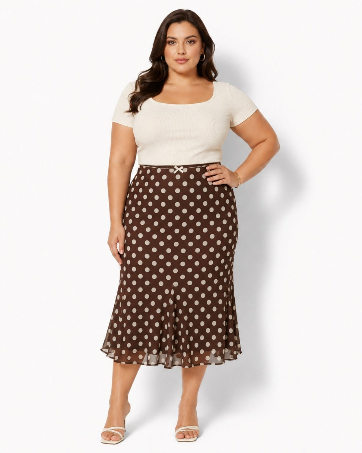 A woman modeling a brown midi skirt with white polka dots and a matching belt, paired with a cream short-sleeve top, against a light gray studio background.