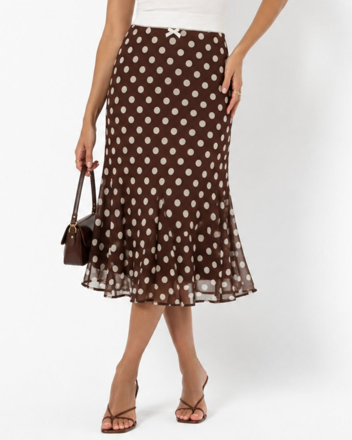 A model wearing a brown midi skirt with white polka dots, styled with a white top, brown strappy heels, and a small brown handbag against a plain white studio background.